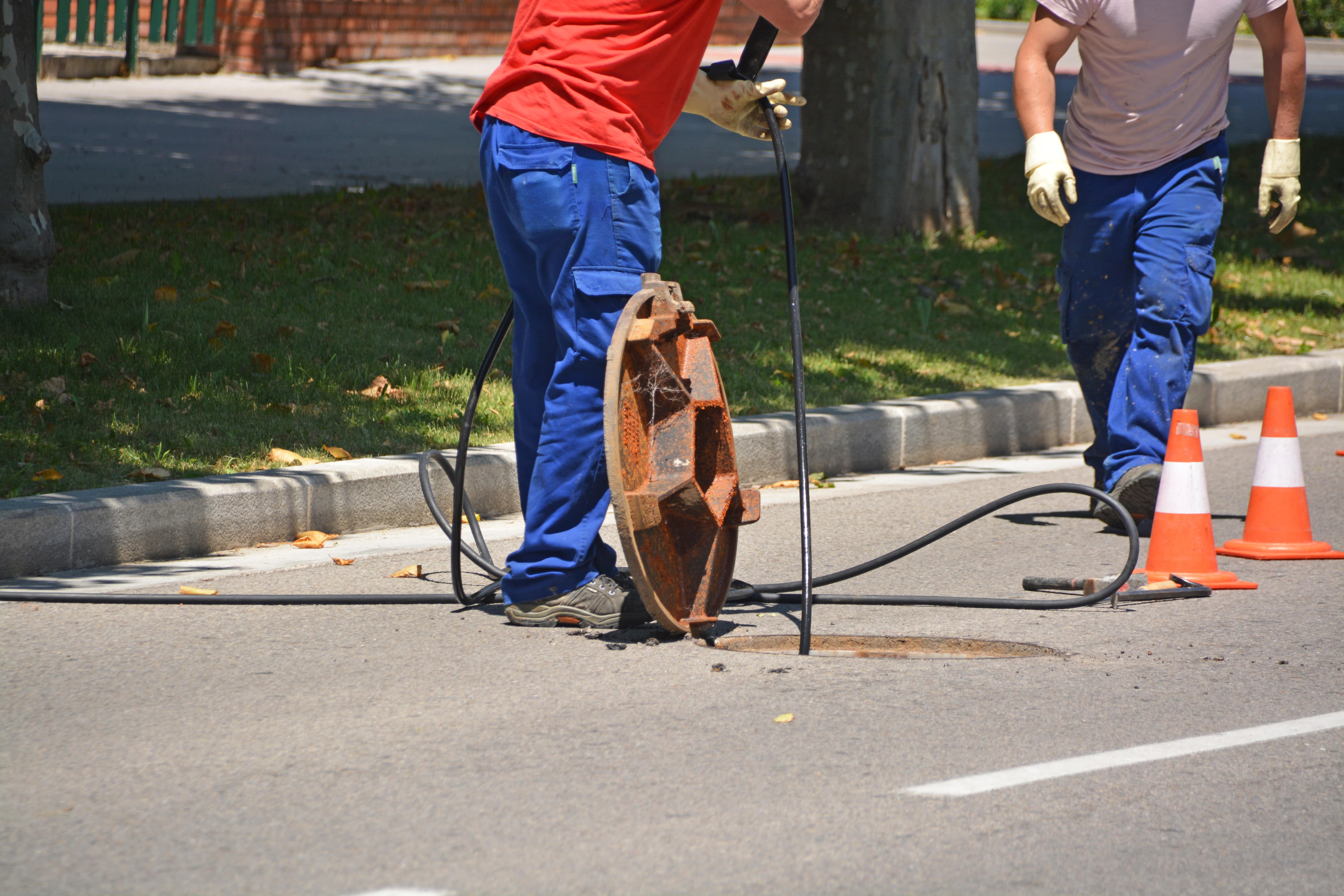 Two workers beside an open manhole in a roadway, with a drain hose fed into the opening