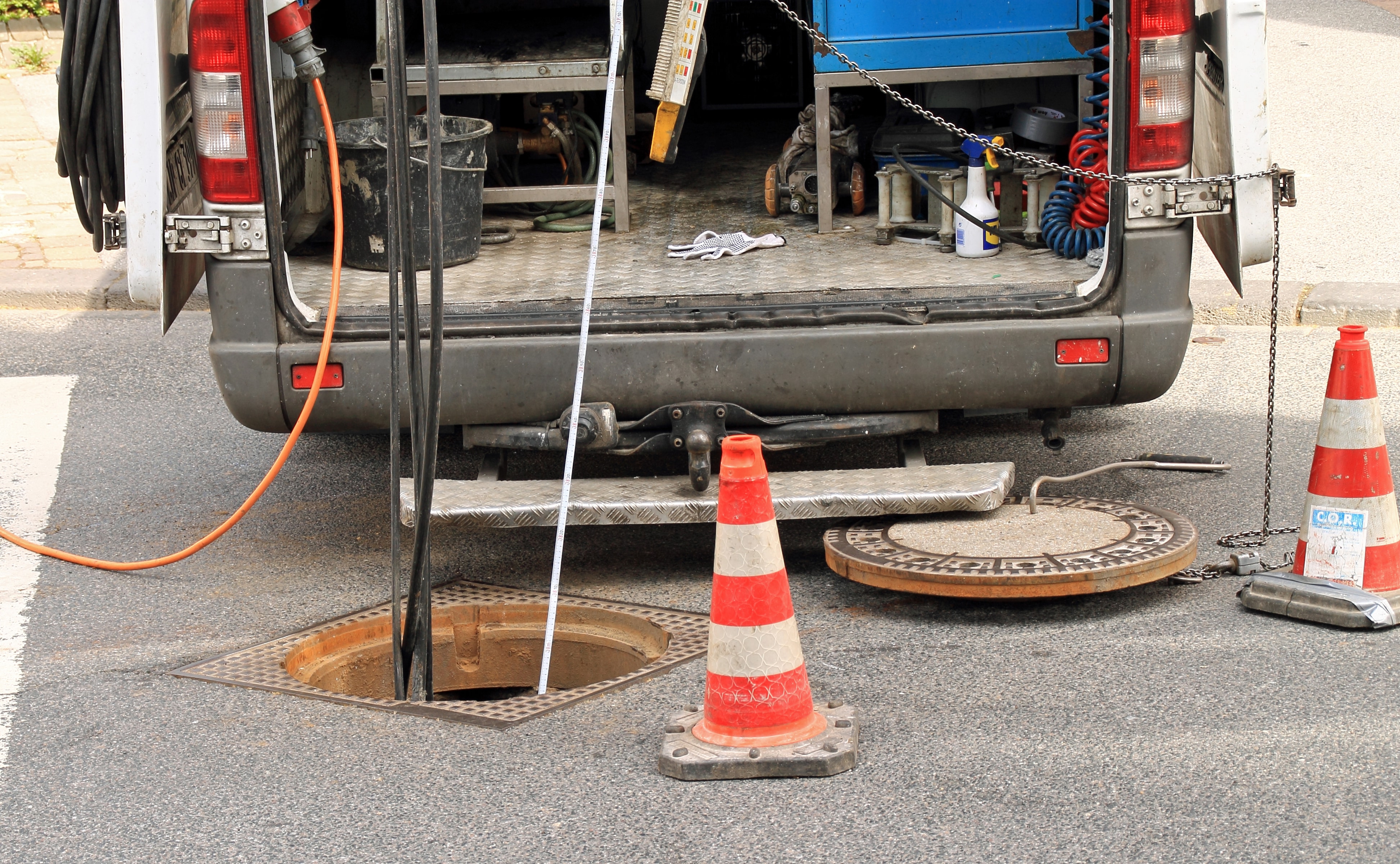 An open service van parked beside a lifted manhole cover, with hoses running down into the drain